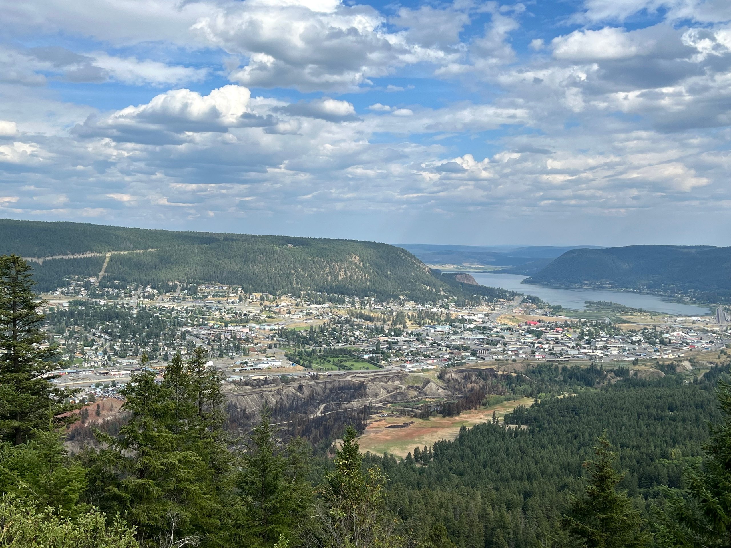 Panoramic view of a forested mountain town, quarry, and winding lake beneath dramatic clouds.