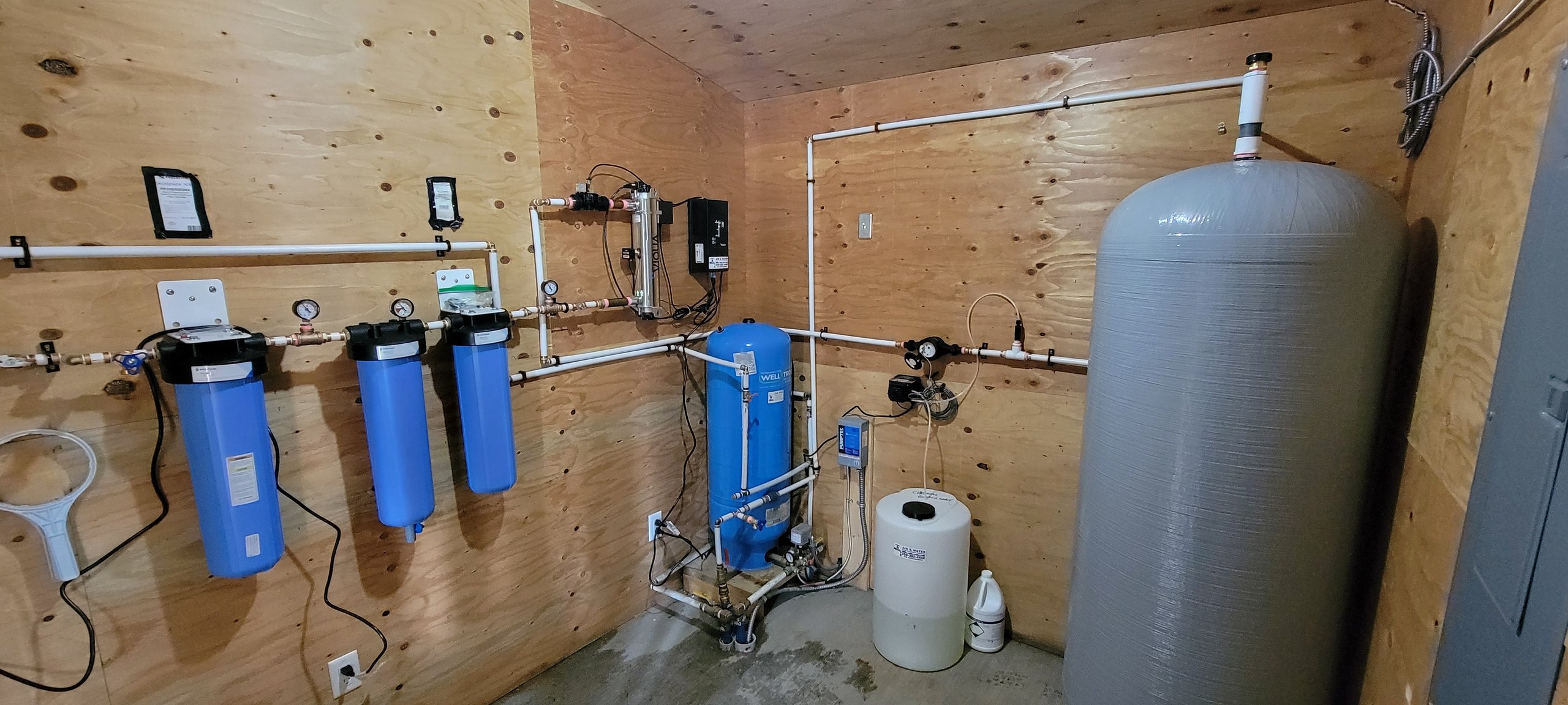 Residential well-water filtration equipment with blue tanks, white piping, gauges, wiring, and a large gray tank in a plywood utility room.